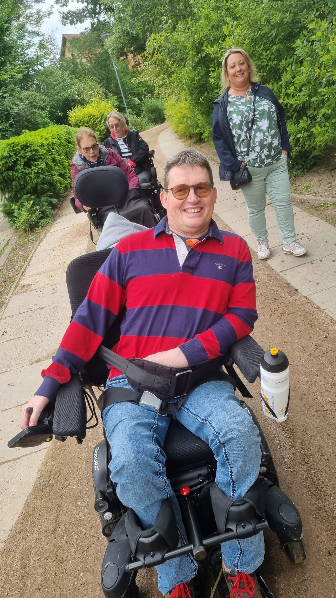 Mann im Rollstuhl lächelt in die Kamera, zwei weitere im Rollstuhl und eine stehende Frau als Assistenzdienst gehen auf Weg hinter ihm.